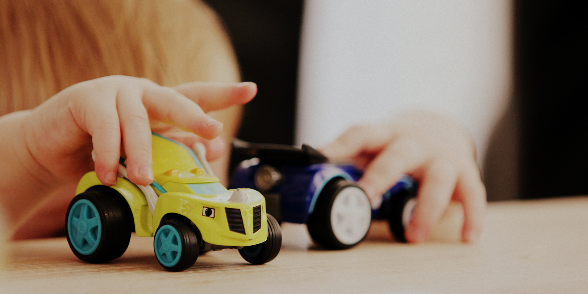 Zdjęcie służy jako tło do cytatu na sliderze głównym. Na zdjęciu dłonie kilkuletniego dziecka bawiącego się dwoma samochodzikami - zabawkami, żółtym i granatowym.